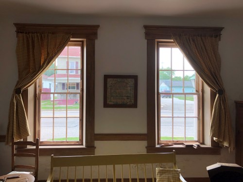 Two windows with checkered curtains, framed by wooden trim, overlooking a street with houses in the background.