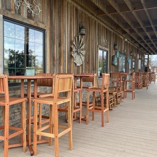 Wooden bar stools and tables line a rustic outdoor patio with stone walls and large windows.