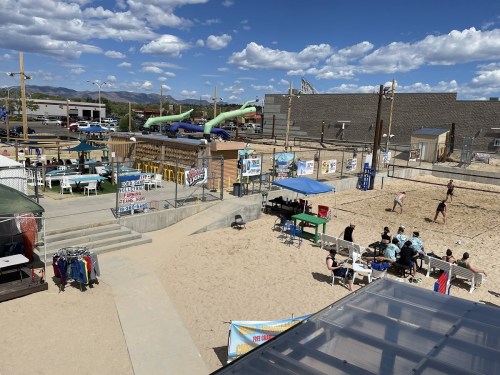 A sunny beach area with volleyball courts, seating, and water slides in the background, surrounded by people enjoying the day.