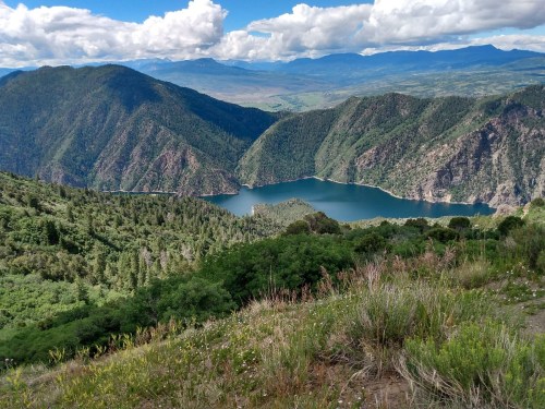A scenic view of a mountainous landscape with a lake surrounded by lush greenery and distant hills under a cloudy sky.