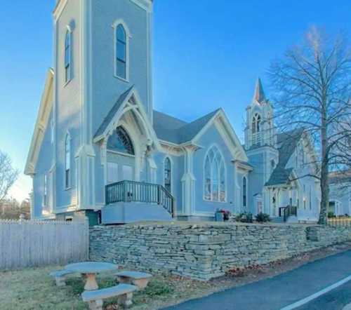 A charming gray church with large windows, surrounded by a stone wall and a picnic table in the foreground.