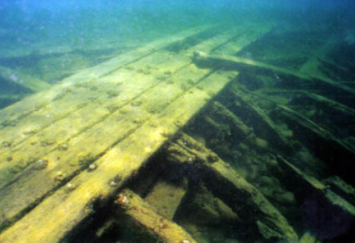 Underwater view of a sunken wooden structure, with beams and planks partially covered by sediment and aquatic life.