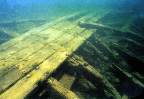 Underwater view of a sunken wooden structure, with beams and planks partially covered by sediment and aquatic life.