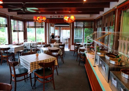 Cozy restaurant interior with checkered tables, wooden chairs, and a buffet area, surrounded by large windows.
