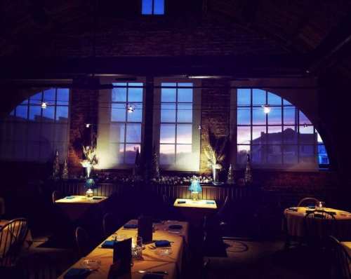 Cozy restaurant interior at dusk, featuring tables set for dining and large windows showcasing a colorful sunset.