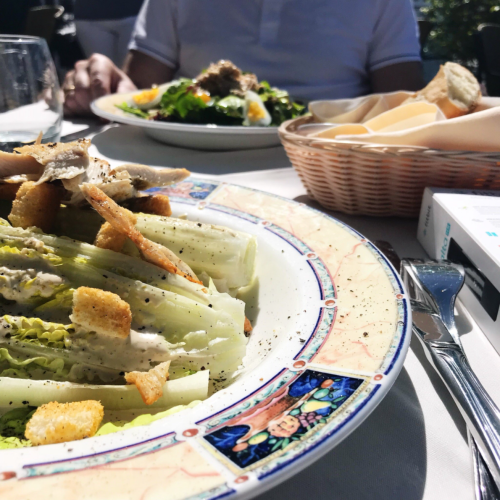 A plate of Caesar salad with croutons and dressing, with a basket of bread and another salad in the background.