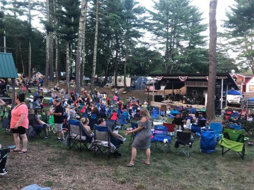 A crowd of people sitting in lawn chairs at an outdoor concert surrounded by trees.
