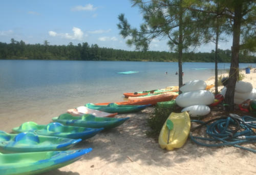Colorful kayaks lined up on a sandy beach by a calm lake, with trees and people enjoying the water in the background.