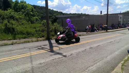 A go-kart with a purple alien figure drives down a street, surrounded by spectators and greenery.