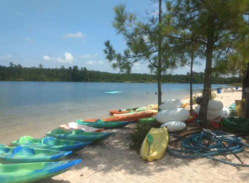 Colorful kayaks and inflatable floats lined up on a sandy beach by a calm lake, surrounded by trees and blue skies.