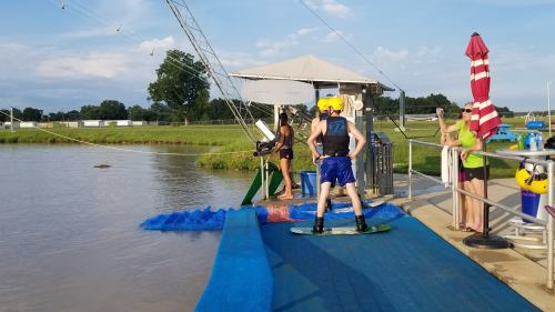 A person prepares to wakeboard at a cable park, while others watch from the dock. Trees and a cloudy sky are in the background.