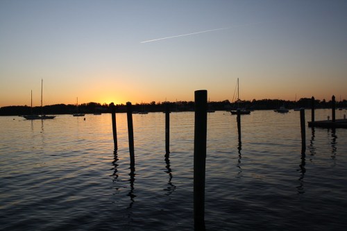 Sunset over a calm water body, with silhouettes of boats and wooden posts reflecting in the water.