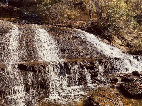 A serene waterfall cascading over rocky steps, surrounded by lush greenery and sunlight filtering through the trees.