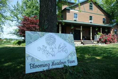 A sign for Blooming Meadows Farm in front of a large house surrounded by greenery and blooming flowers.