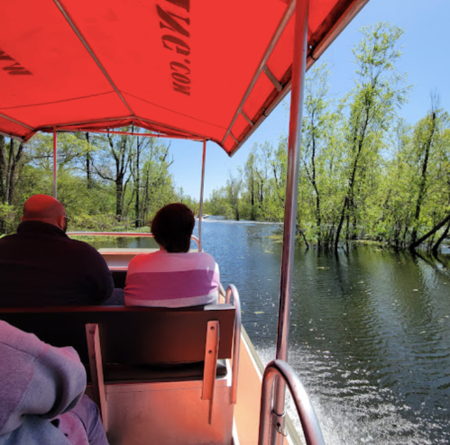 Two people sit under a red canopy on a boat, surrounded by lush greenery and calm waters on a sunny day.