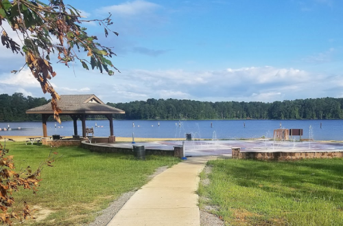 A scenic view of a lake with a gazebo, fountains, and a pathway leading to the water under a blue sky.