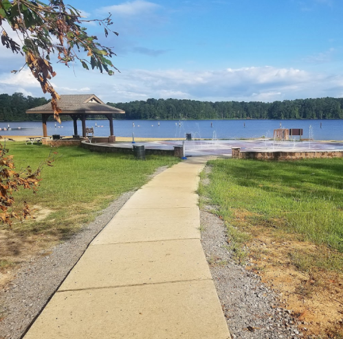 A paved path leads to a lake with a gazebo and fountains, surrounded by green grass and trees under a blue sky.