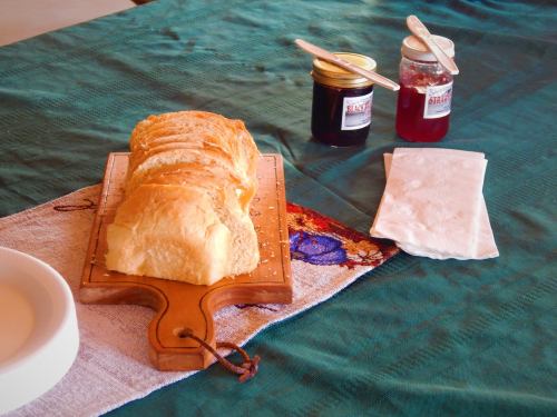 Sliced bread on a wooden board next to two jars of jam and a napkin on a green tablecloth.