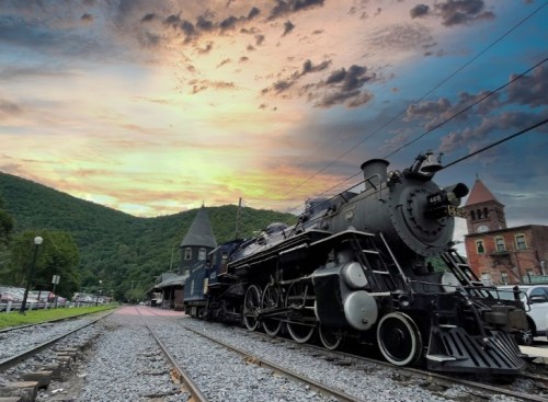 A vintage steam locomotive parked at a station, with mountains and a colorful sunset in the background.
