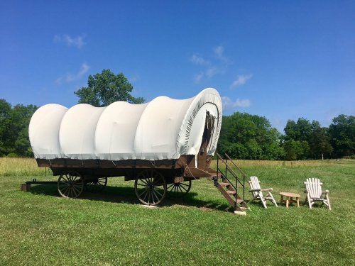 A covered wagon on wheels sits on grass, with two chairs and a small table nearby under a clear blue sky.