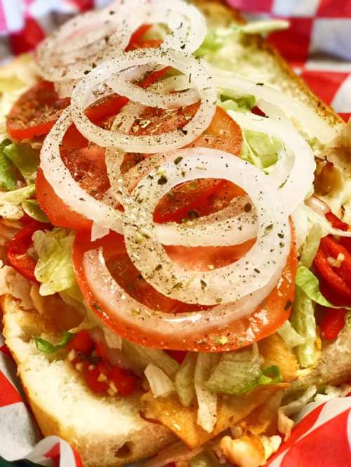 Close-up of a sandwich topped with lettuce, tomatoes, onions, and spices on toasted bread, served in a red checkered basket.