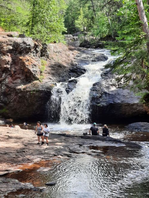 A scenic waterfall cascades over rocks, with people enjoying the water and surrounding greenery.