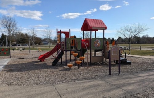Colorful playground with slides and climbing structures, surrounded by gravel and open green space under a blue sky.