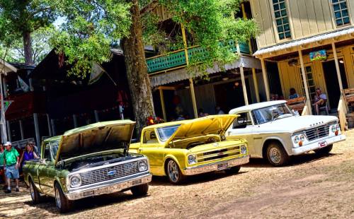 Three vintage cars with hoods open parked under trees near a rustic building, with people in the background.