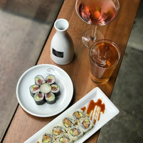 A wooden table with sushi rolls, a sake bottle, and two drinks: a cocktail and a beer.