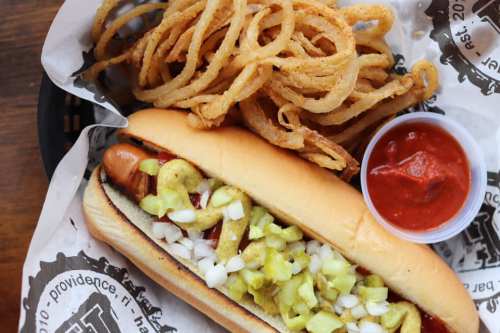 A hot dog topped with onions and pickles, served with crispy onion rings and a side of sauce in a paper-lined basket.