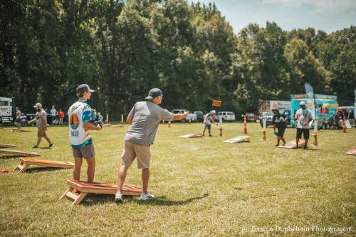 Two people playing cornhole on a grassy field, with several boards and spectators in the background.