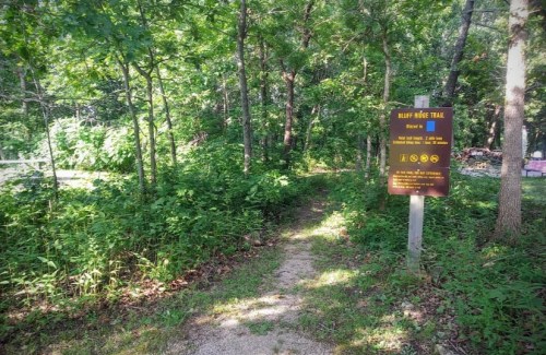 A trail sign for "Hilly Ridge Trail" surrounded by lush greenery and trees, indicating the path ahead.