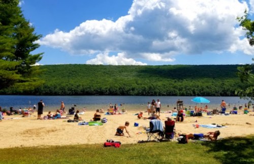 A sunny beach scene with people relaxing on the sand and by the water, surrounded by green hills and fluffy clouds.