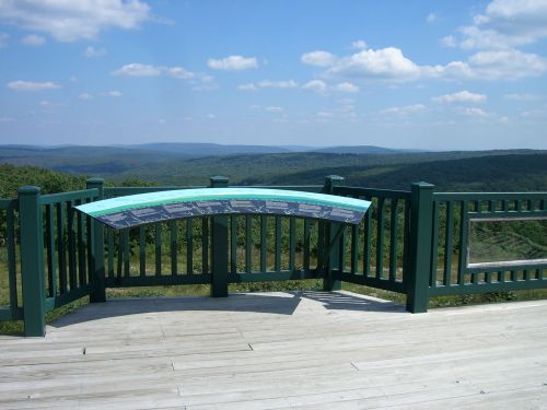 A scenic overlook with a curved informational panel, surrounded by green railings and a panoramic view of rolling hills.