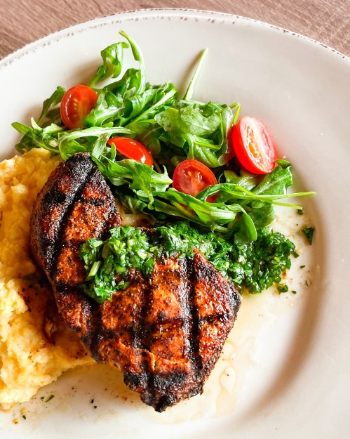 Grilled fish fillet topped with green sauce, served with creamy polenta and a side of arugula and cherry tomatoes.