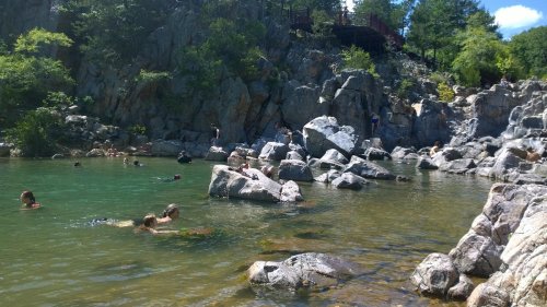 People swimming and relaxing in a natural pool surrounded by rocks and greenery on a sunny day.