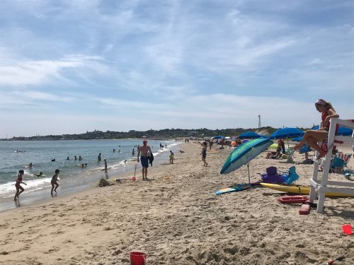 A sunny beach scene with people swimming, playing, and relaxing under umbrellas, while a lifeguard watches from a chair.