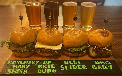 A wooden platter with four gourmet sliders labeled: Rosemary Baby Swiss, Da Brie, Beet Slider, and BBQ Baby, alongside four beers.