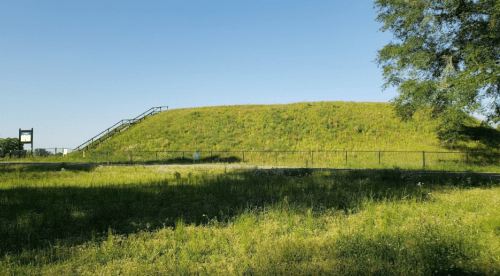 A grassy mound with a staircase on one side, surrounded by a green field and clear blue sky.