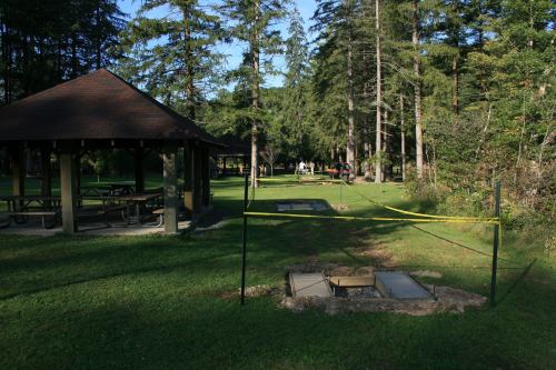 A park scene with a gazebo, picnic tables, and a fenced area in a grassy landscape surrounded by trees.
