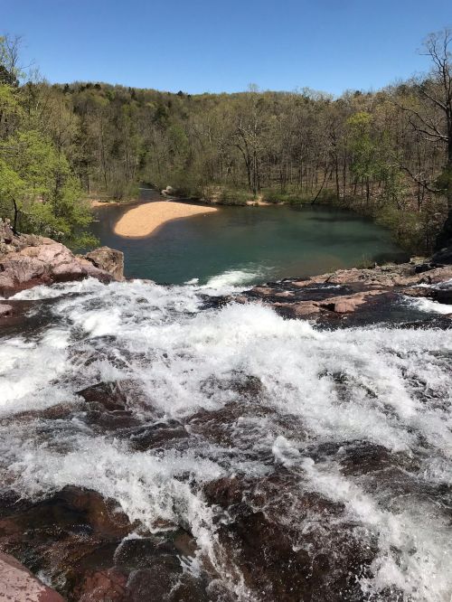 A scenic view of a waterfall cascading into a calm river, surrounded by lush greenery and a sandy beach in the distance.