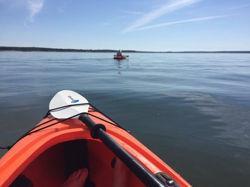 A view from an orange kayak, with another kayak in the distance on a calm, sunny lake.