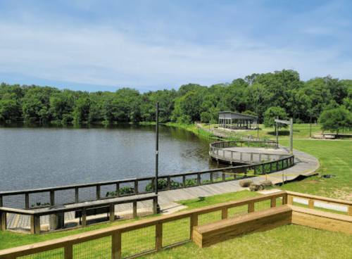 A serene lake surrounded by greenery, featuring wooden walkways and a gazebo under a clear blue sky.
