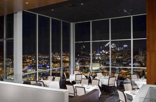 Elegant restaurant interior with white tablecloths and a stunning city skyline view at night through large windows.