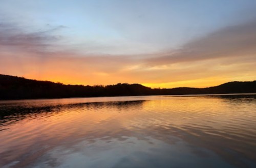 A serene lake at sunset, reflecting vibrant orange and purple hues against a calm water surface and distant hills.