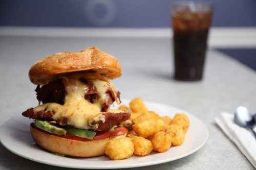 A close-up of a cheesy chicken sandwich with lettuce and tomato, served with tater tots and a glass of soda in the background.