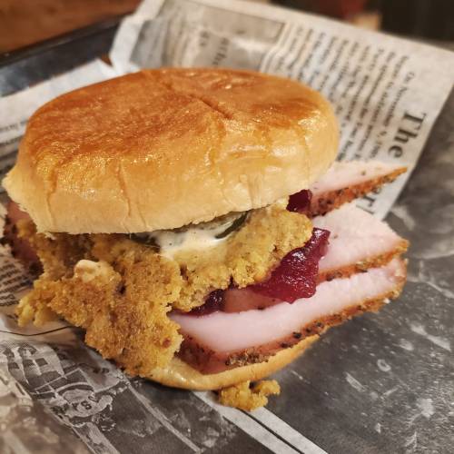 A sandwich featuring sliced turkey, stuffing, and cranberry sauce on a toasted bun, wrapped in newspaper.