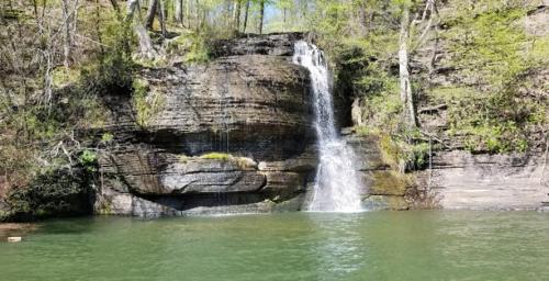 A serene waterfall cascading over rocky cliffs into a calm green pool, surrounded by lush trees and greenery.