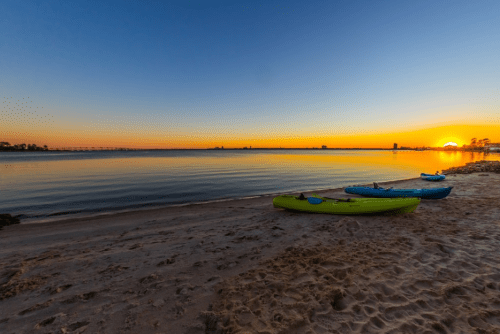 Two kayaks sit on a sandy beach at sunset, with calm waters and a vibrant sky reflecting the colors of dusk.