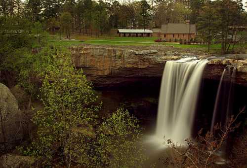 A serene waterfall cascades over a rocky ledge, surrounded by lush greenery and a rustic building in the background.